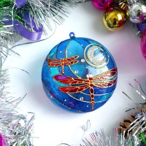 May include: A blue glass Christmas ornament with a painted design of two dragonflies and a crescent moon. The dragonflies are red, gold, and blue.