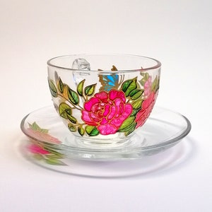 Puede incluir: Un juego de taza y platillo de vidrio con diseños florales pintados a mano. La taza presenta rosas rosadas, hojas verdes y una mariposa azul. El platillo tiene un diseño floral a juego.