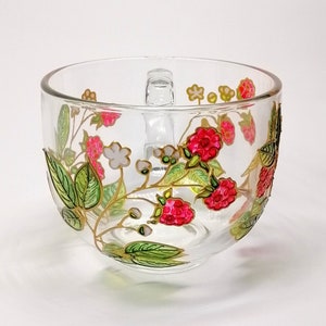Peut inclure: Une tasse en verre transparent avec un bord doré et une anse. La tasse est décorée d'un motif peint à la main de framboises et de feuilles dans des tons de vert, rose et blanc.