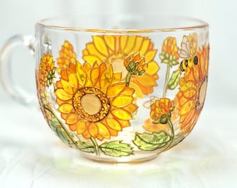May include: Clear glass teacup with a handle, decorated with hand-painted sunflowers in shades of yellow and orange. The design includes green leaves and a small bee. The teacup is perfect for enjoying a warm beverage.