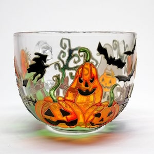 Może przedstawiać: Przezroczysta szklana miska z motywem Halloween. Miska jest ozdobiona ręcznie malowanymi dyniami, nietoperzami i czarownicą na miotle. Dynie są pomarańczowe i zielone z czarnymi oczami i ustami.
