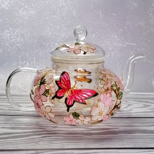 Può includere: Una teiera di vetro trasparente con una farfalla rosa e un motivo floreale. La teiera ha un infusore rimovibile e un coperchio con un pomello.