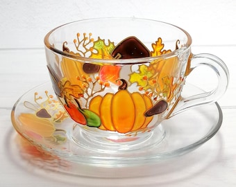 May include: A glass teacup and saucer set with a hand-painted autumn design featuring pumpkins, leaves, and berries. The cup and saucer are clear glass with a delicate gold trim.