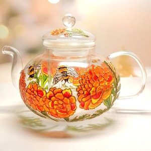 以下が含まれることがあります： オレンジと黄色の花と蜂で飾られた、透明なガラス製のティーポット。蓋と取っ手があります。