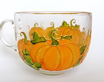 May include: A clear glass mug with a hand-painted design of orange pumpkins with green leaves and stems. The pumpkins are arranged in a cluster and have a textured appearance.