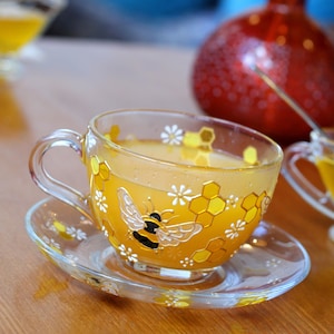 Può includere: Una tazza da tè di vetro con un design di api e favi, riempita di un liquido giallo. La tazza da tè è su un piattino abbinato.