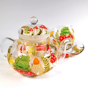Puede incluir: Un juego de tetera y taza de té de vidrio pintado a mano con diseños de hongos y ranas. La tetera tiene un cuerpo de vidrio transparente con tapa y colador. La taza tiene un cuerpo de vidrio transparente con asa. Ambas piezas están decoradas con ranas verdes, hongos rojos y blancos y hojas verdes.