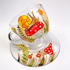 May include: A glass teacup and saucer set with a hand-painted design of a snail on a mushroom with red and white polka dots. The cup and saucer are decorated with green leaves and gold accents.