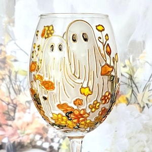 Op de afbeelding: Een helder wijnglas met een geschilderd ontwerp met twee vriendelijke geesten en herfstkleurige bloemen. De geesten zijn wit met zwarte ogen en de bloemen zijn oranje, geel en bruin. Het glas heeft een lange steel en een ronde voet.
