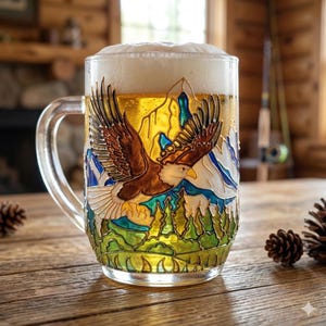 Könnte beinhalten: Ein klares Bierglas, gefüllt mit Bier und Schaum. Das Glas hat ein detailliertes, farbenfrohes Design eines fliegenden Adlers über Bergen und Bäumen. Der Griff ist klar, und das Glas steht auf einer Holzoberfläche.