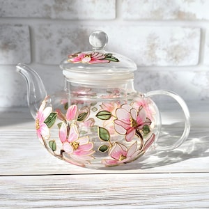 May include: A clear glass teapot with a lid and handle, adorned with pink and gold floral designs and green leaves. The teapot sits on a white wooden surface, with a white brick wall in the background.
