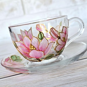 Puede incluir: Un juego de taza y platillo de té de vidrio con flores de magnolia pintadas a mano en rosa y dorado. La taza y el platillo son de vidrio transparente con un delicado diseño floral.