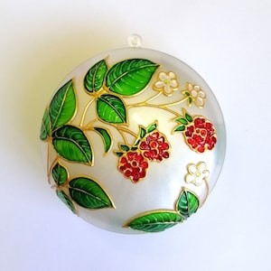 Op de afbeelding: Een witte, ronde ornament met een gouden rand en een geschilderd ontwerp van groene bladeren, rode frambozen en witte bloemen.