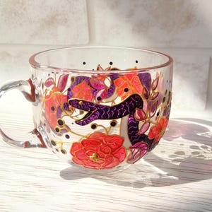 May include: A clear glass mug with a hand-painted design featuring a purple snake winding through pink and red flowers. The snake is outlined in gold, and the flowers have gold accents.