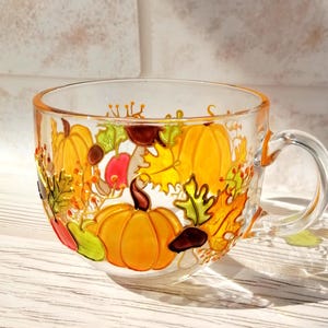 Pode incluir: Uma caneca de vidro transparente com alça, pintada à mão com um design outonal que apresenta abóboras, folhas e bolotas em tons de laranja, amarelo, verde e marrom.
