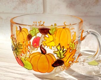May include: A clear glass mug with a handle, hand-painted with an autumnal design featuring pumpkins, leaves, and acorns in shades of orange, yellow, green, and brown.