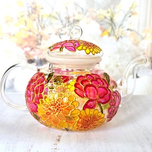 May include: A clear glass teapot with a lid and handle, decorated with hand-painted floral patterns in pink, yellow, and orange. The flowers are outlined in gold. This decorative teapot is designed for brewing tea and adds a touch of elegance.