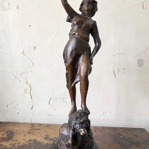 Peut inclure: Statue en bronze d'une femme tenant un objet floral en l'air. La figure est debout sur une base avec une créature ressemblant à un lion. La statue a une patine brun foncé et est montée sur une base en bois noir.