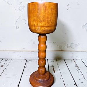 May include: A wooden pedestal bowl with a round base and a stem of stacked spheres. The bowl is a light brown color and sits on a white-painted wooden surface. The background is a white wall with texture.