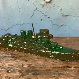 Op de afbeelding: Een vintage groen speelgoed schip met afbladderende witte verf. De speelgoedboot heeft een gedetailleerd ontwerp met meerdere schoorstenen en een hoge mast. Het schip staat op een houten oppervlak.