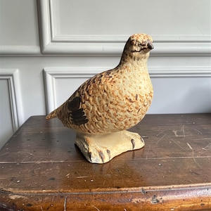 May include: A ceramic bird figurine with a detailed, textured surface. The bird is primarily tan and brown with black accents on its wings. It is perched on a light-colored, rectangular base, and is a decorative item.