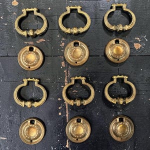 May include: A collection of twelve brass furniture hardware pieces arranged in four rows. The set includes six oval-shaped drop pulls with decorative detailing and six circular backplates featuring a central recessed circle and a single mounting hole.
