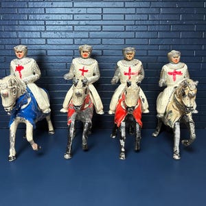 May include: Four toy knights on horseback. The knights wear white tunics with red crosses and helmets. The horses are painted in various colors, including white, gray, and black, with blue and red accents. The figures are set against a dark blue brick wall.