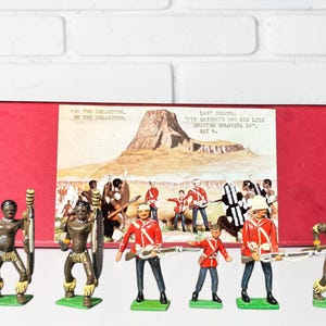 Puede incluir: Un juego de juguetes vintage con figuras en miniatura de soldados y guerreros. Las figuras están pintadas en rojo, negro y dorado. El juego incluye una caja roja con una ilustración de una escena de batalla y texto.