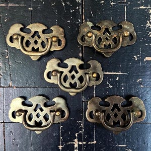 May include: Five matching brass drawer pulls with a decorative openwork backplate and a swinging bail handle. Each piece features a symmetrical, intricate cutout design with a warm, aged metallic finish and two mounting points for attachment.