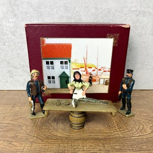 以下が含まれることがあります： 漁師、魚屋、兵士のミニチュアフィギュアをフィーチャーしたヴィンテージジオラマ。シーンには、魚のテーブル、樽、ボートと建物のある沿岸の町の額入り写真が含まれています。フレームはバーガンディです。