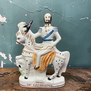 May include: A ceramic figurine of the Duke of Cambridge on horseback. The figure is dressed in a white uniform with blue accents and gold details. The horse is white with blue polka dots and a tan saddle. The base reads "Duke of Cambridge."
