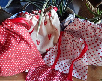 May include: Four drawstring bags with red hearts on a white background. The bags are made of fabric and have red ribbon ties.