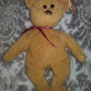 May include: A golden-brown teddy bear with black eyes and a brown nose, featuring a burgundy ribbon and a red tag. This plush toy is a classic teddy bear, a popular children's toy.