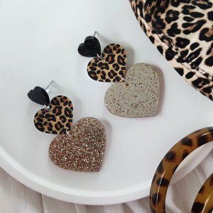 Könnte beinhalten: Ein Paar Ohrringe mit einem mehrschichtigen Herzdesign. Das obere Herz ist ein schwarzer und brauner Leopardenprint, das mittlere Herz ist ein solides schwarzes Herz und das untere Herz ist ein goldenes Glitzerherz.