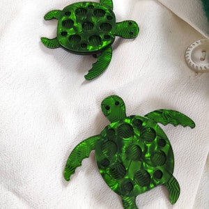 Puede incluir: Dos broches en forma de tortuga verde con un acabado texturizado y marmoleado. Las tortugas tienen un patrón circular en relieve en sus caparazones.