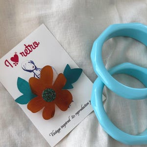 May include: A retro-themed image featuring a flower brooch and two light blue bangles. The brooch has an orange flower with a green glitter center and teal leaves. A card with the text "I retro" is also present.