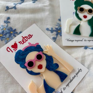 May include: Two retro-style brooches featuring a woman with blue hair, pink sunglasses, and a red lip. One brooch has green hair and sunglasses. The brooches are on white cards with the text "I love retro" and "Vintage inspired & reproduction jewelry."