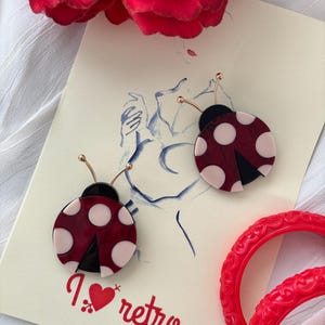 May include: A pair of red and white ladybug earrings with black spots. The earrings are on a white card with the text "I love retro" in red.