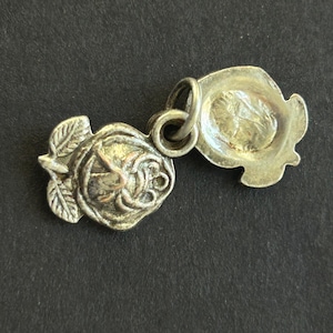 May include: Two silver-toned charms connected by a small ring. One charm is a detailed rose with leaves, the other is a hat-shaped charm. The charms are likely vintage or antique, with a tarnished appearance.