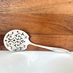 May include: A silver serving spoon with a decorative, openwork design on the head. The handle is long and slender, with a floral pattern near the end. The spoon is set against a wooden background.