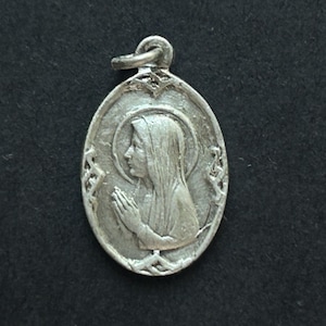 May include: Oval silver pendant featuring a detailed relief of a woman in profile with hands clasped in prayer. The pendant has a loop at the top for a chain and decorative edging. The background is a dark, textured surface.