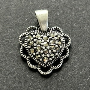 May include: A silver heart-shaped pendant with a detailed filigree design. The heart is filled with small, sparkling, gray-black marcasite stones.