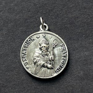 May include: A silver-colored St. Patrick pendant with the words "ST. PATRICK" and "PRAY FOR US" encircling an image of St. Patrick holding a staff. The pendant has a loop at the top for a chain.