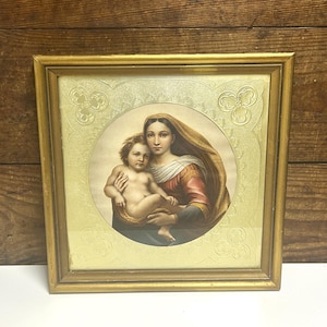 May include: A framed religious print depicting a mother and child. The image is set within a gold-coloured frame. The artwork shows a woman in a red and gold robe holding a child. The background is a light yellow colour.