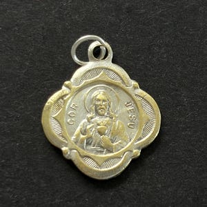 May include: A two-tone metal pendant featuring a central relief of Jesus with a halo and a heart, surrounded by a quatrefoil frame. The text COR JESU is embossed on the sides. The pendant includes a small silver-colored jump ring at the top.