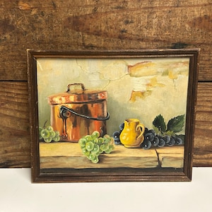 May include: A framed still life painting depicting a copper pot, a yellow jug, and bunches of green and purple grapes. The artwork has a vintage feel, with a wooden frame and a neutral backdrop. The painting is likely an oil painting.