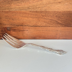 May include: A silver fork with an ornate handle and four tines. The fork is lying on a white surface with a wooden background.