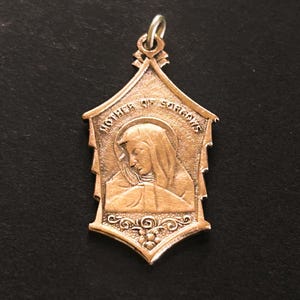 May include: A bronze-toned metal pendant featuring a relief image of a veiled figure with a halo. The text MOTHER OF SORROWS is inscribed in an arch above the figure. The pendant has a pointed top and bottom with decorative scrollwork at the base.