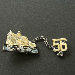 May include: A vintage pin featuring a gold-toned building labeled "Franciscan Monastery Washington D.C." connected by a chain to a gold-toned number "56". The pin has a blue border.