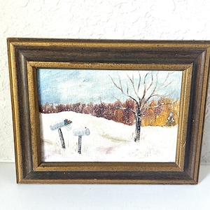 May include: Framed winter landscape painting featuring a snowy scene with two mailboxes and a bare tree. The artwork has a gold-toned frame and depicts a snow-covered field and a forest in the background.